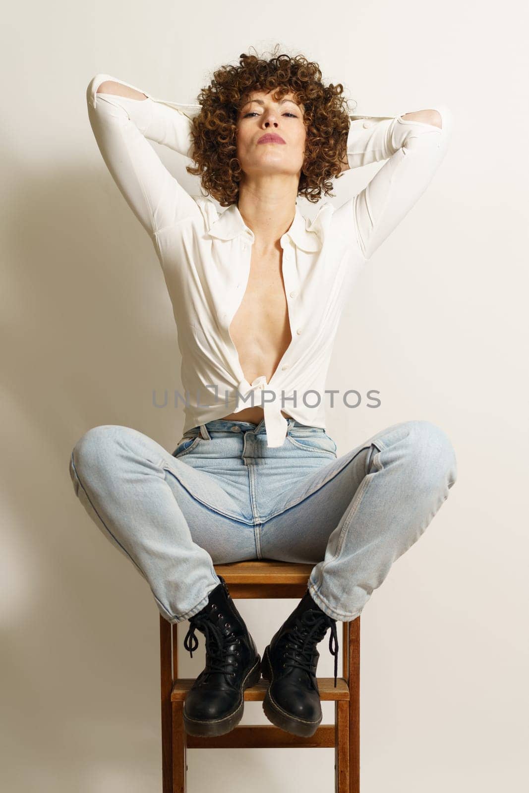 Full length of bossy female model with red hair, in stylish clothes resting on wooden stool with hands on head and staring at camera against white wall