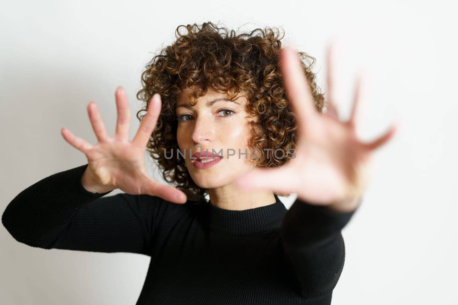 Attractive young female model, with curly hair looking at camera and showing stop gesture with full open palms and fingers against gray background in blurring unseen side illuminating lights