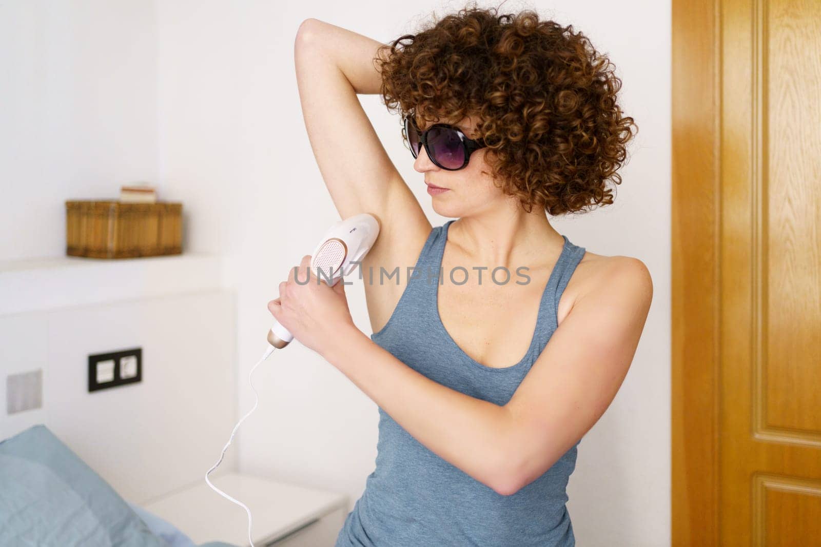 Concentrated young female in gray top and protective goggles with curly hair removing hair from armpit with pulsed light laser during beauty routine at home