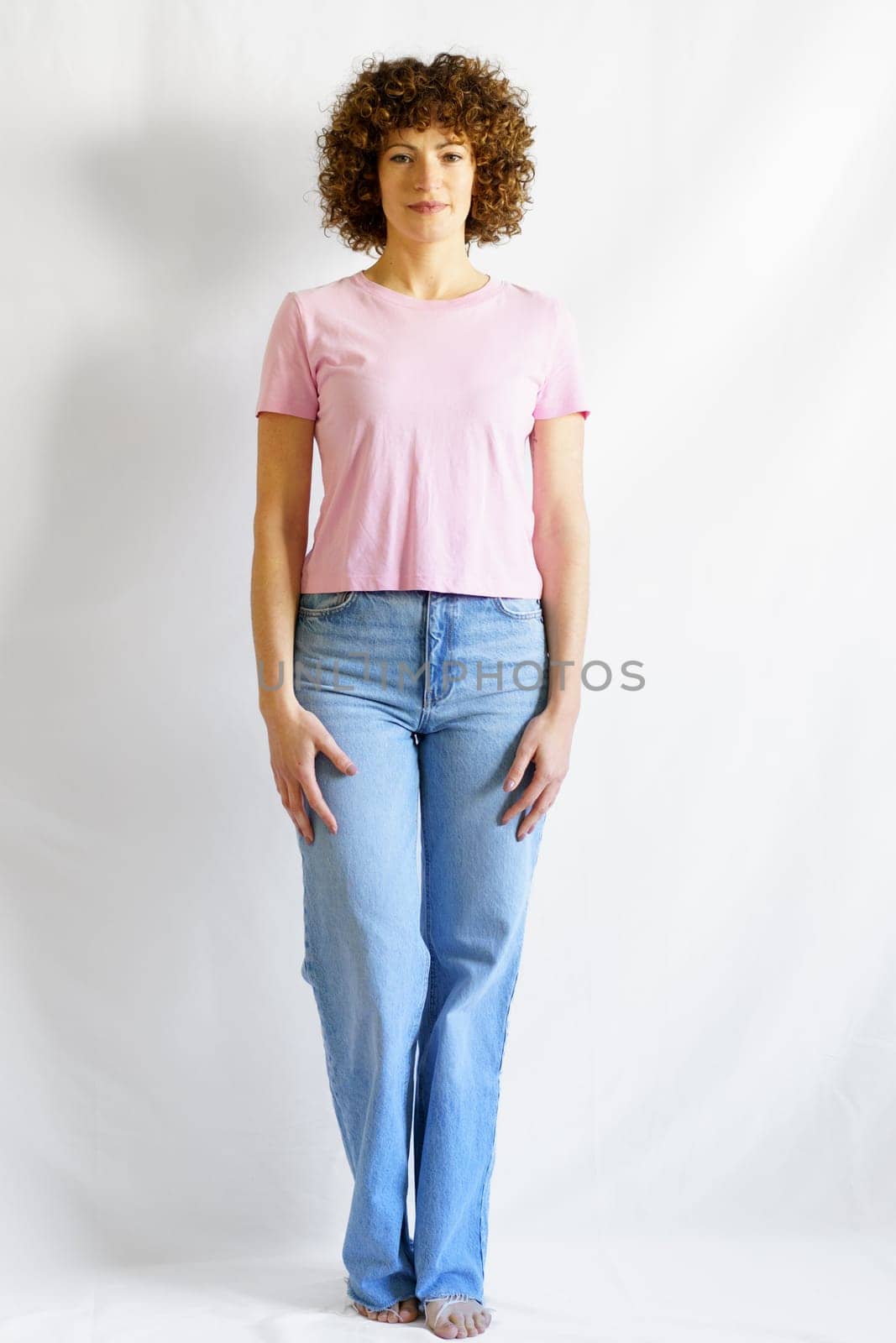 Full body of curly haired female in jeans and pink t-shirt leaning on white wall looking at camera