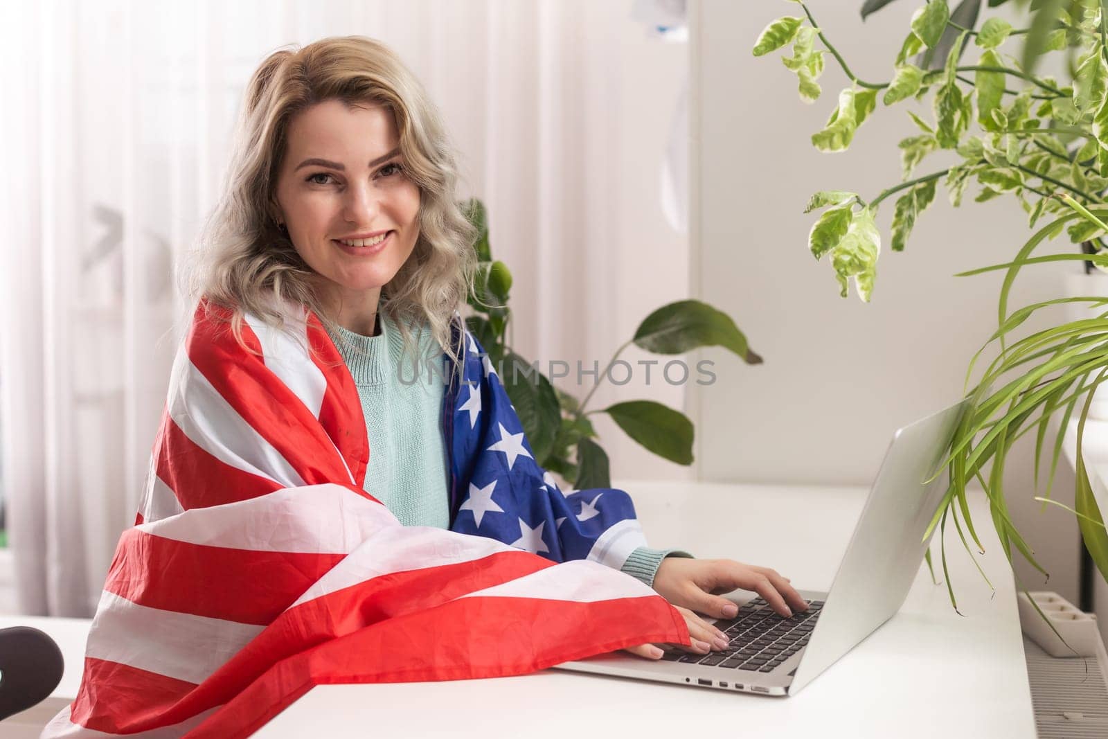 Happy woman employee sitting wrapped in USA flag, shouting for joy in office workplace, celebrating labor day or US Independence day
