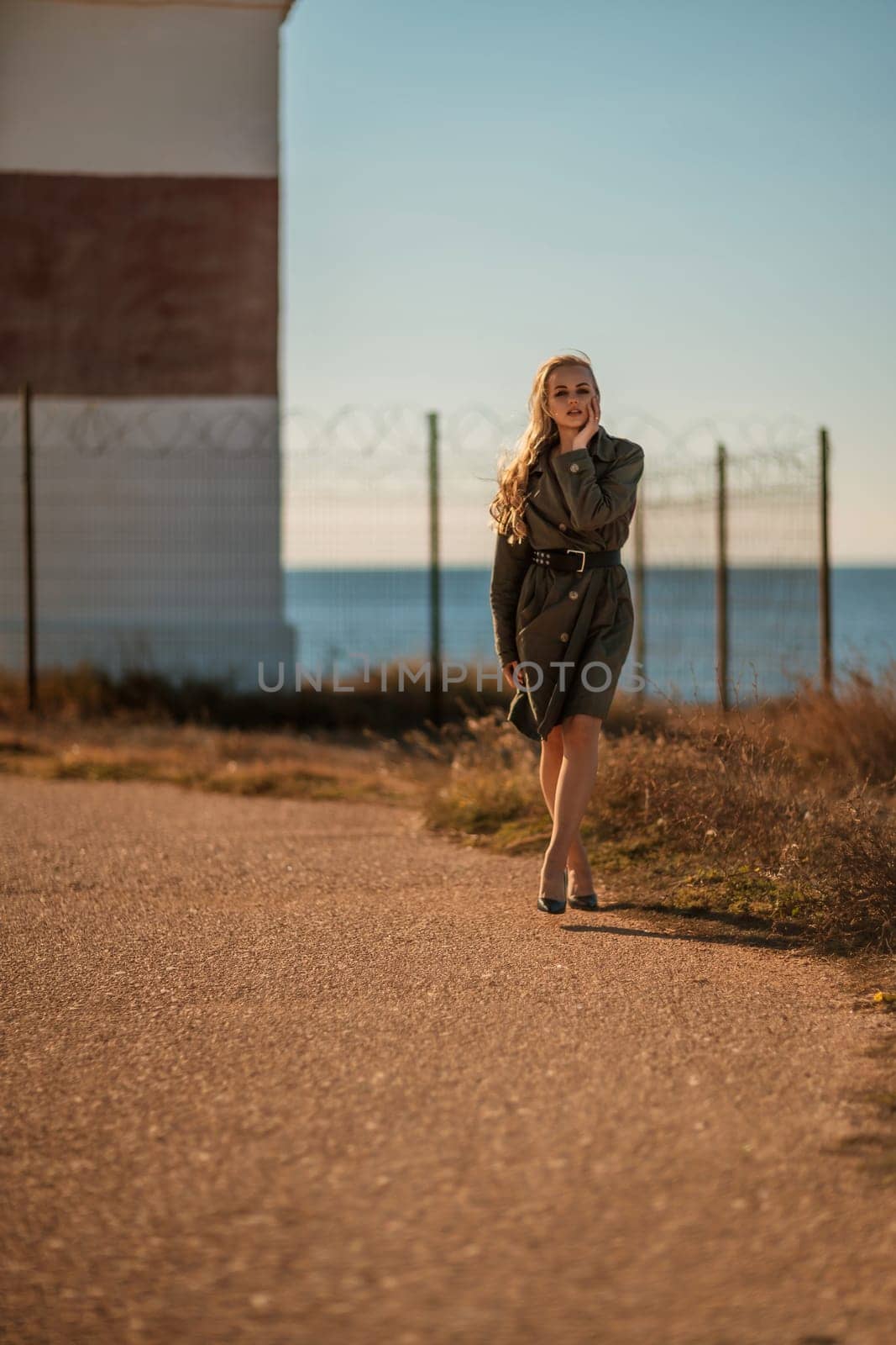 Portrait blonde sea cape. A calm young blonde in a khaki raincoat stands on the seashore against the backdrop of a lighthouse. by Matiunina