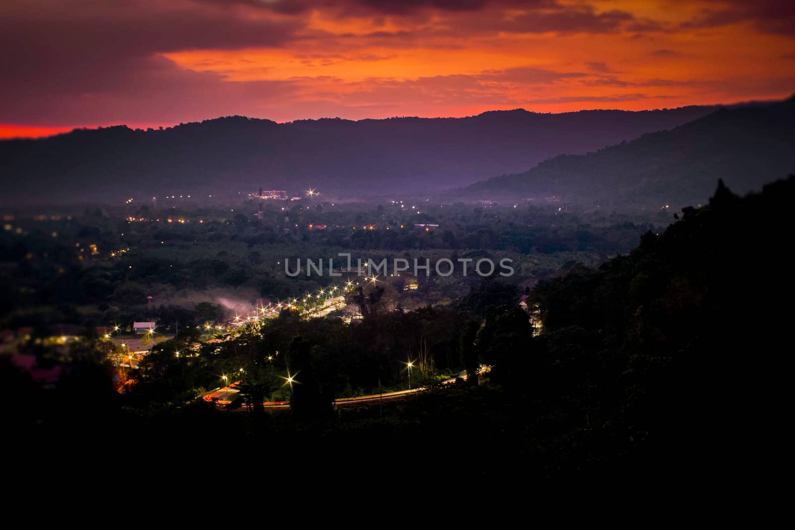 Landscape with alpine mountain valley, low clouds, forest, purple sky with stars, city illumination at sunset. Aerial.