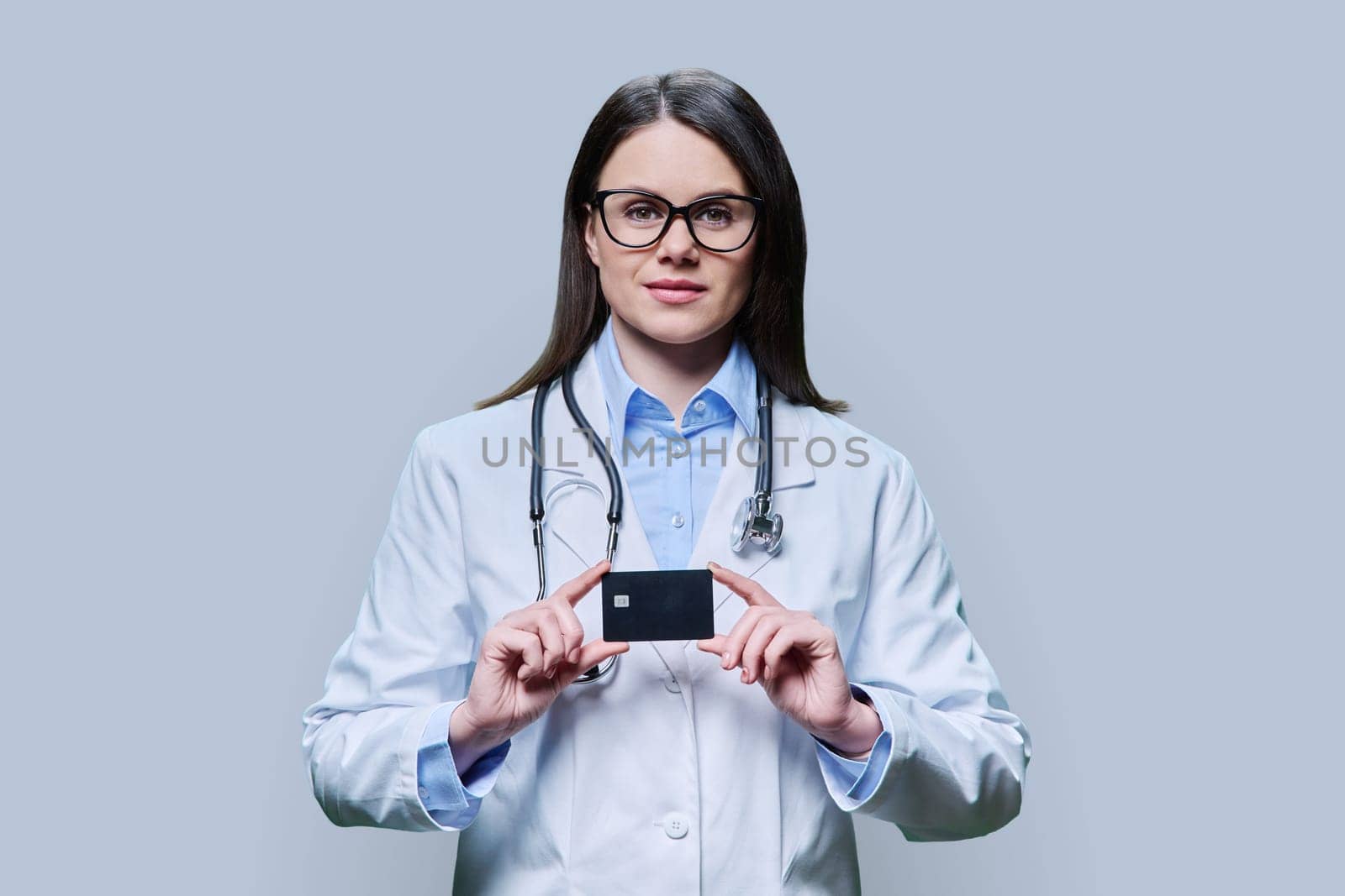 Young woman doctor holding credit card on light gray studio background. Friendly female looking in camera. Insurance, healthcare, medicine, credit card service concept