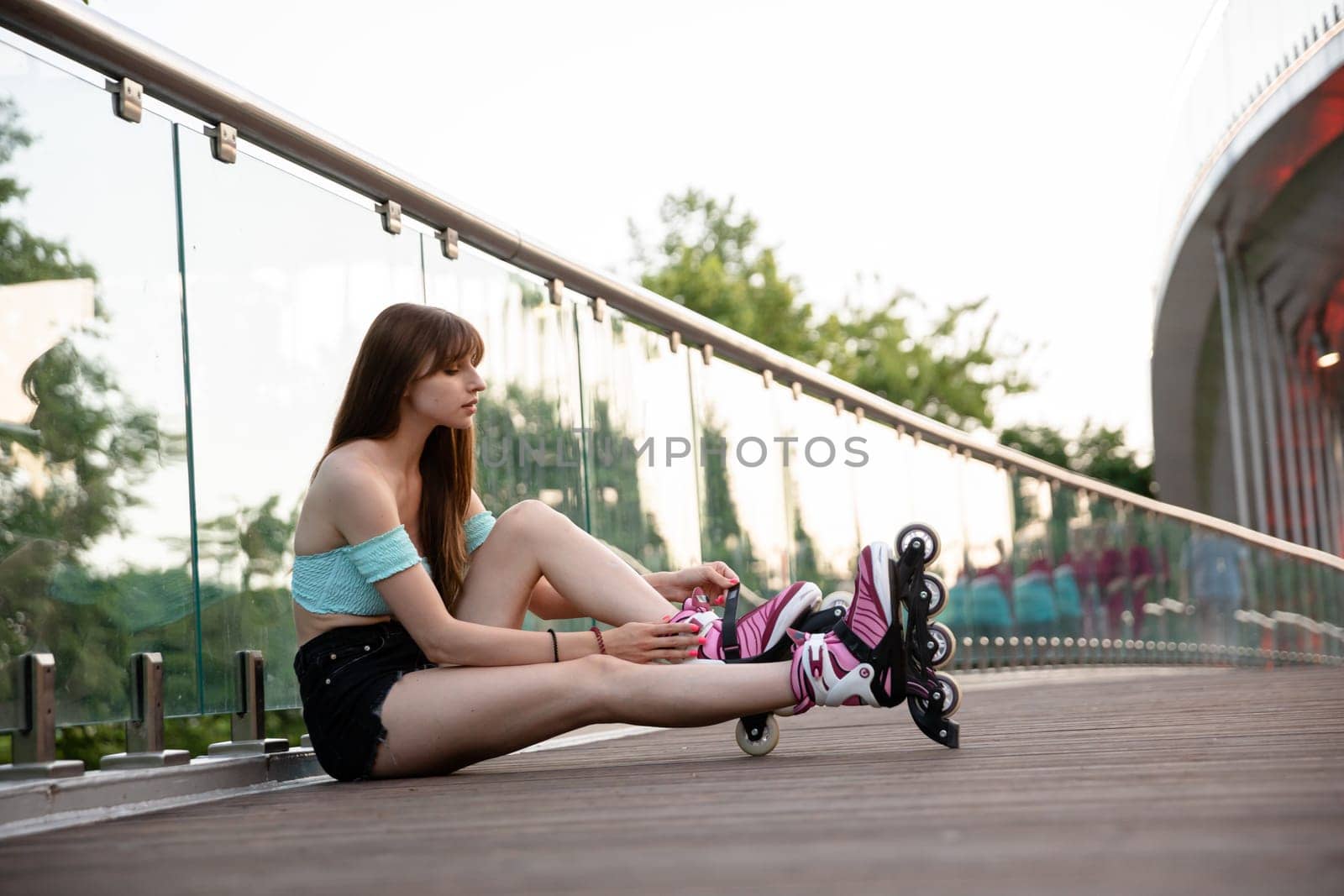 The girl is sitting on a wooden bridge. She has a glass balustrade behind her back. In the background you can see green treetops. The young woman is wearing summer clothes, a blue top and black shorts.