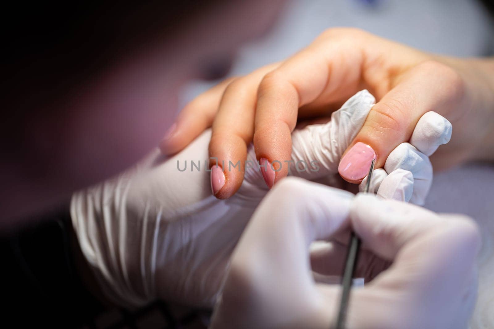 Blurred profile of a woman in the foreground. A beautician paints a client's nails with pink polish. Performing a manicure using the hybrid method. Making an appointment at a beauty salon.