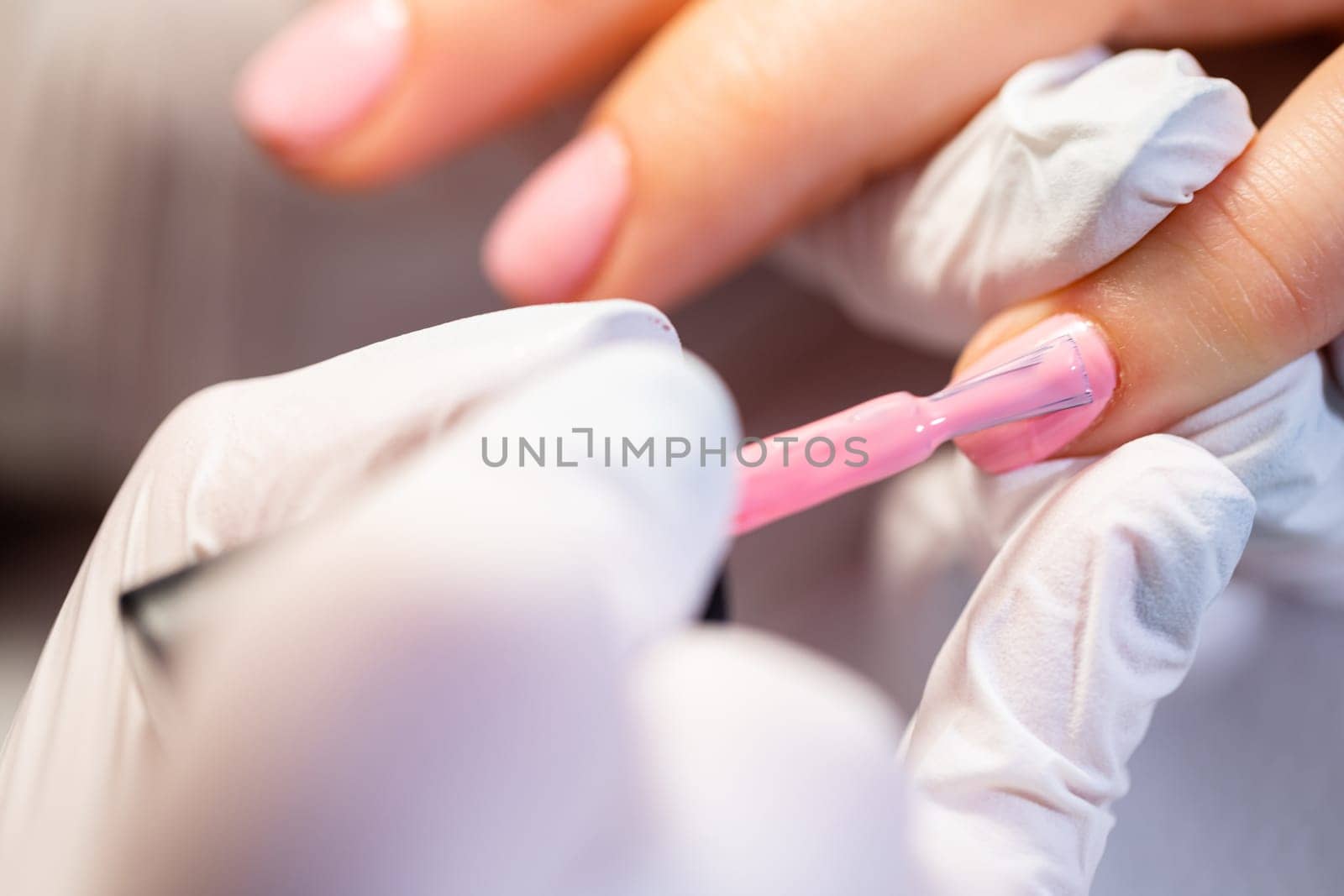 Blurred foreground. Large close-up on the hands of a working manicurist. The beautician is performing a hybrid manicure. The professional paints the woman's nails pink.