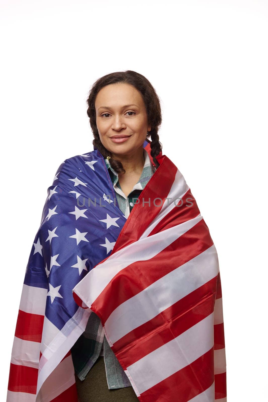 Proud multinational woman wrapped in the flag of the United States of America, looking at camera on isolated white background, proud to be American citizen. Copy ad space. July 4th. Independence Day