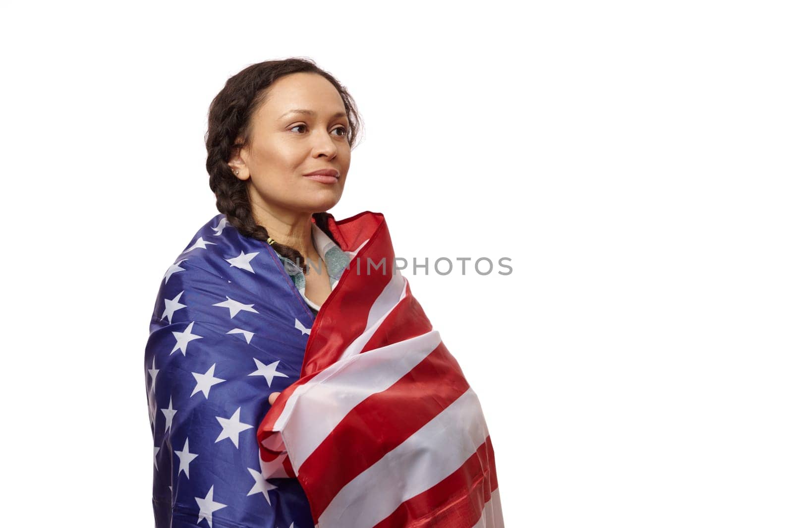Proud multinational woman wrapped in the flag of the United States of America, looking away on an isolated white background, proud to be an American citizen. Copy ad space. July 4th. Independence Day
