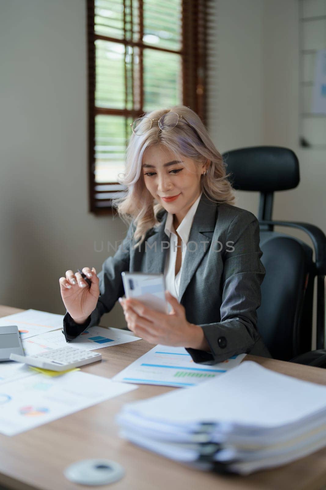 Portrait of a young Asian woman showing a smiling face as she uses her phone, computer and financial documents on her desk in the early morning hours.