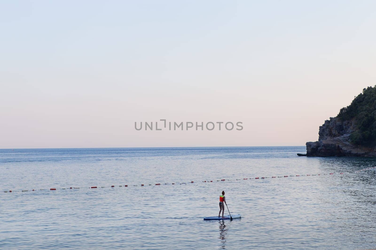 A girl with a paddle in the sea stands on a sapboard at sunrise. Active rest on the sea. by sfinks