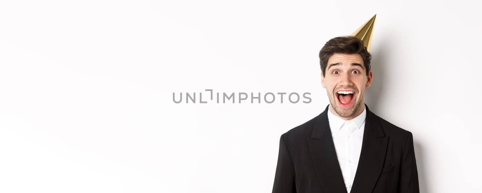 Close-up of excited handsome guy in trendy suit and party hat, looking amazed, celebrating winter holidays, standing against white background.