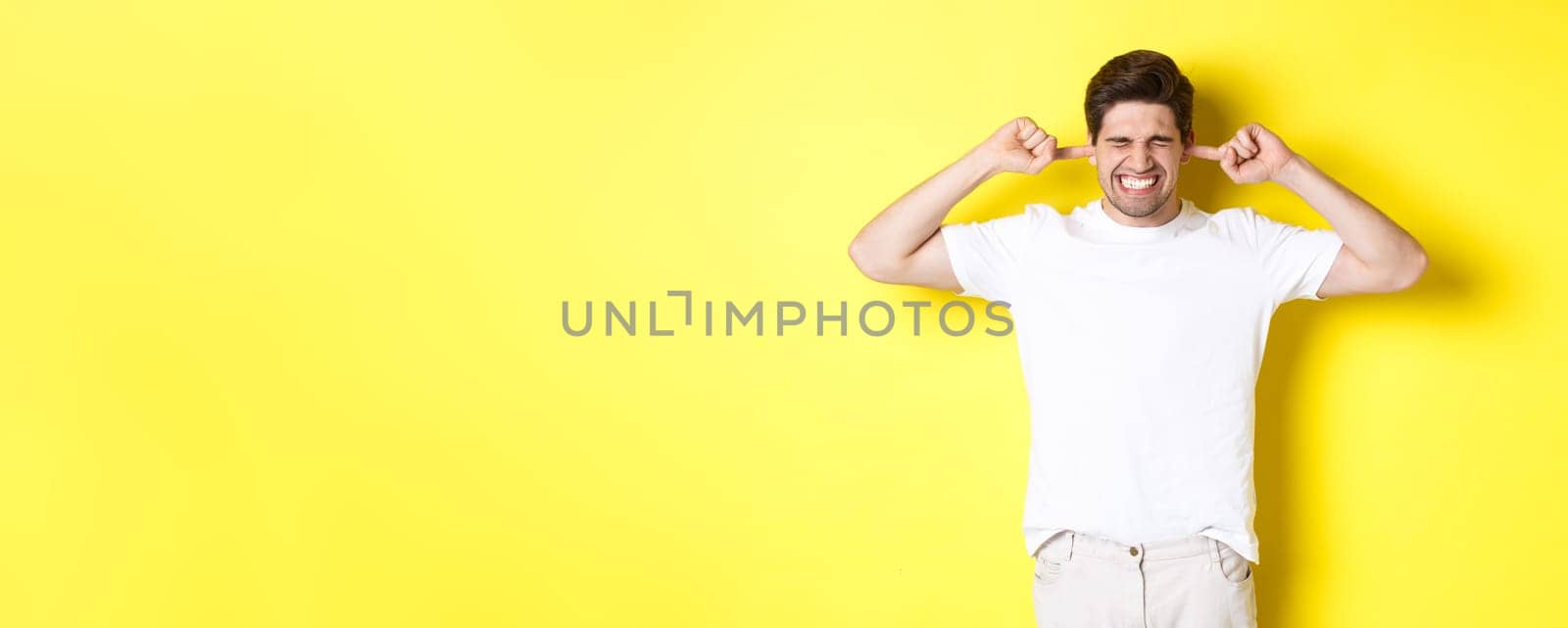 Annoyed man grimacing and shutting ears, complaining on loud noise, standing against yellow background. Copy space