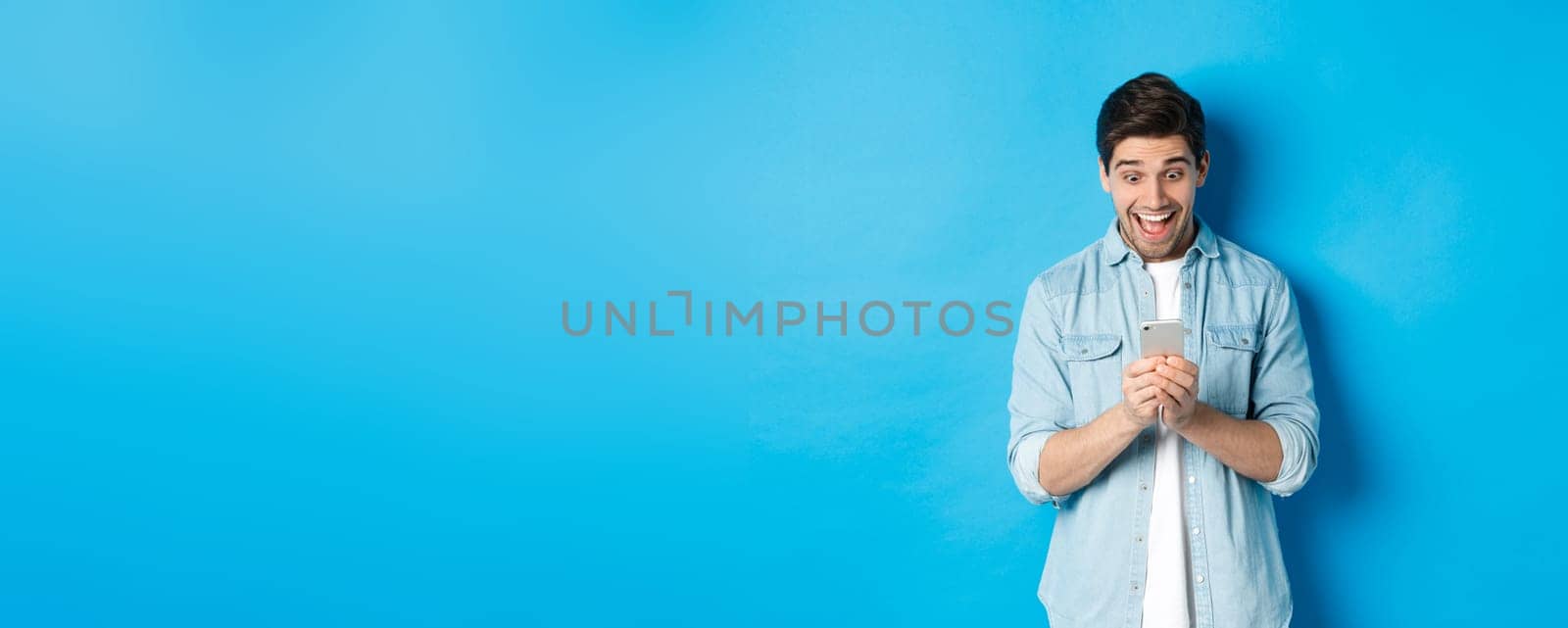 Image of excited man smiling while looking at mobile phone, shopping online on smartphone, standing against blue background.