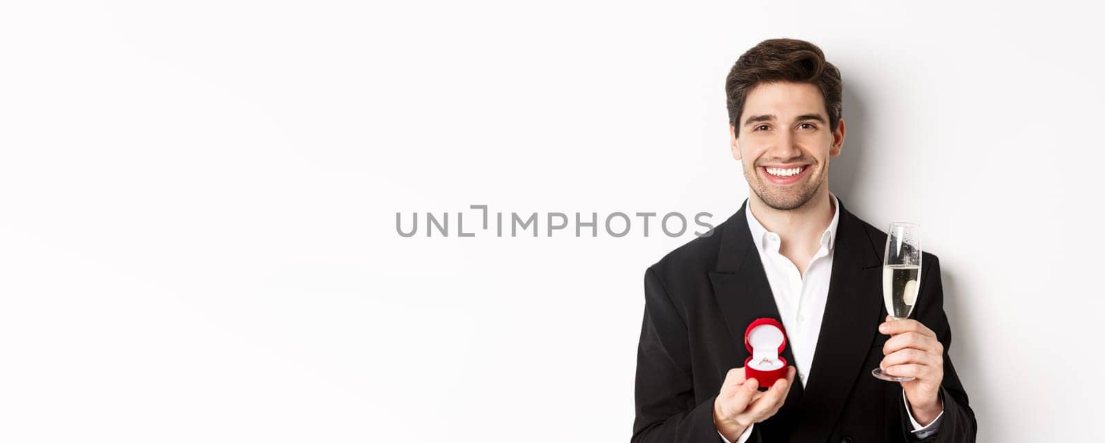 Close-up of handsome man in suit, making a proposal, giving engagement ring and raising glass of champagne, standing against white background.