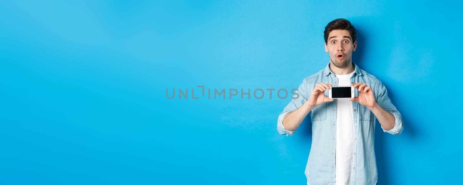 Surprised guy showing mobile phone screen and looking impressed, standing over blue background by Benzoix