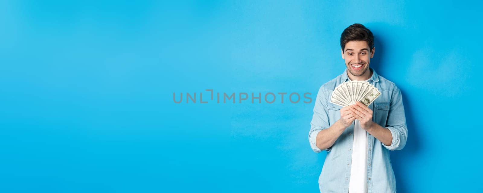 Excited successful man counting money, looking satisfied at cash and smiling, standing over blue background by Benzoix