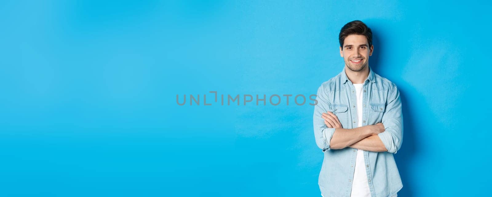 Handsome guy in casual clothes standing with arms crossed and confident smile against blue background.