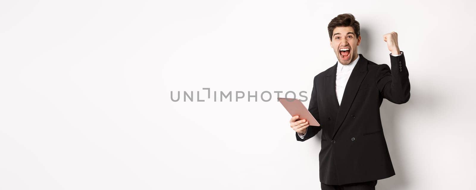 Portrait of handsome successful businessman in black suit, achieve goal, holding digital tablet and saying yes, standing against white background and rejoicing.