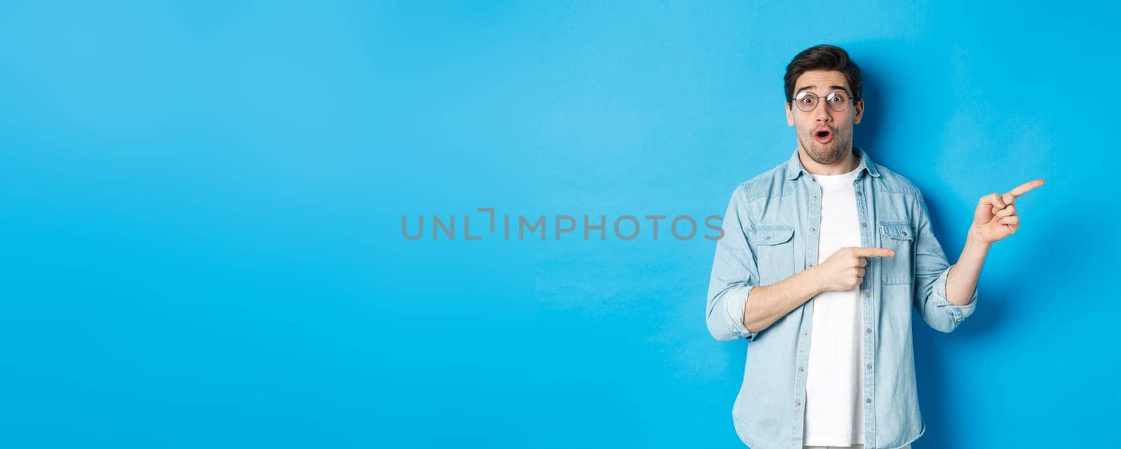 Surprised man in glasses pointing right at copy space, showing promo offer on blue background, standing over blue background by Benzoix