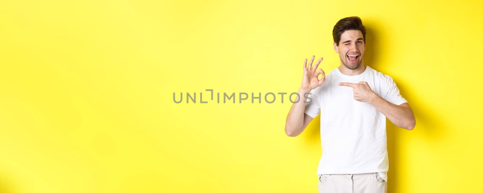 Image of handsome young man approve something, showing okay sign and winking, standing against yellow background.