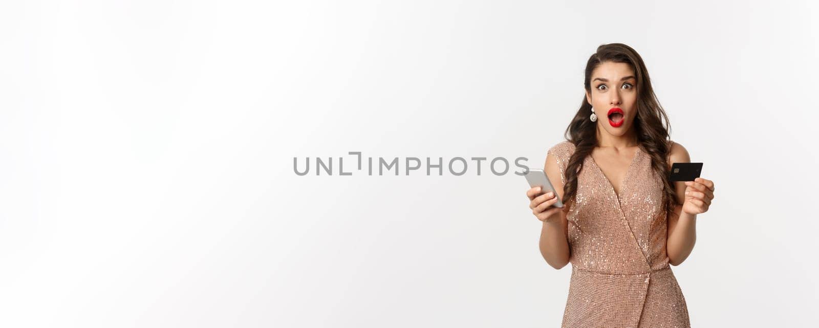 Online shopping and holidays concept. Surprised woman in glamour dress, buying in internet with credit card and mobile phone, looking amazed, white background.
