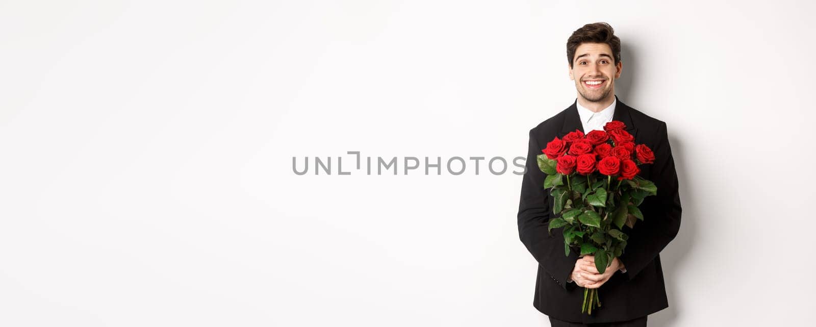 Image of handsome man in black suit, holding bouquet of roses and smiling, standing against white background by Benzoix