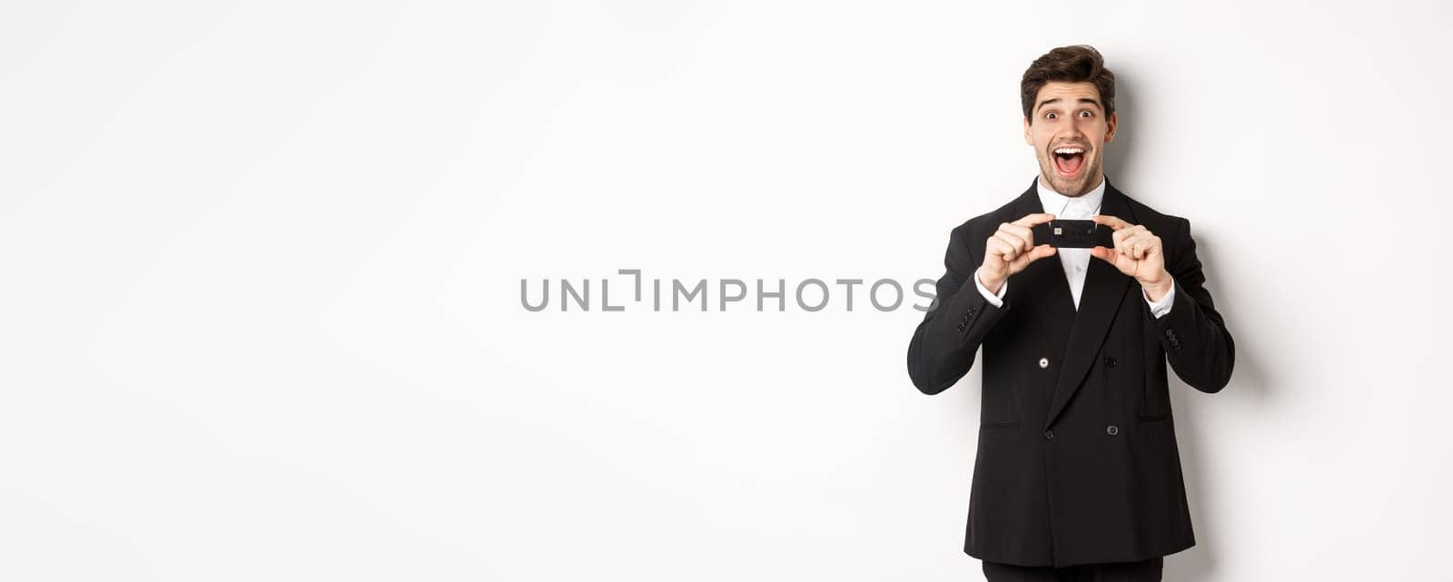 Portrait of amazed handsome man in black suit, showing credit card and recommending bank, standing over white background by Benzoix