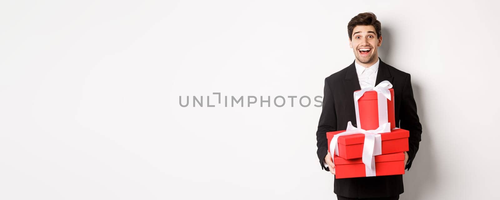Concept of holidays, relationship and celebration. Handsome man in black suit bringing presents at new year party, holding gifts and smiling amused, standing against white background by Benzoix