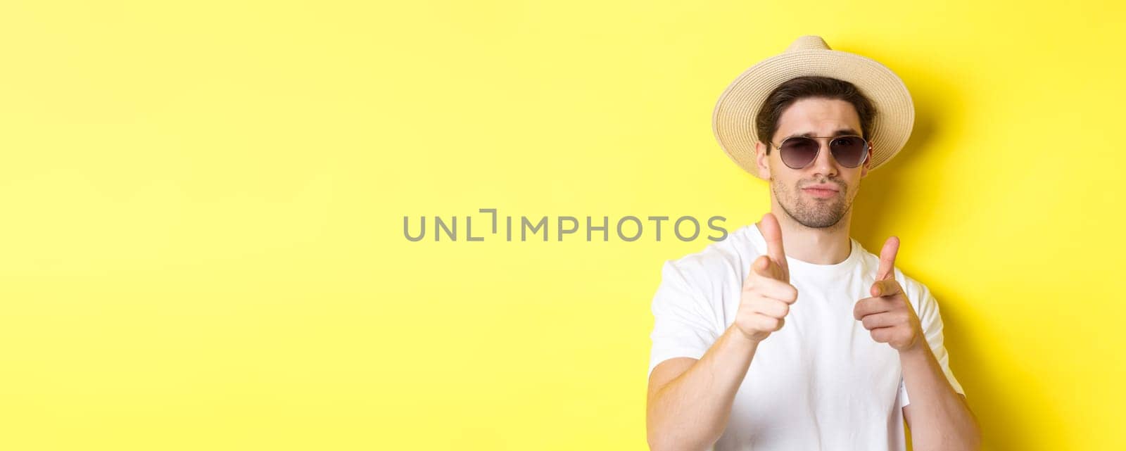 Concept of tourism and vacation. Close-up of cool guy in summer hat and sunglasses pointing finger guns at camera, standing over yellow background by Benzoix