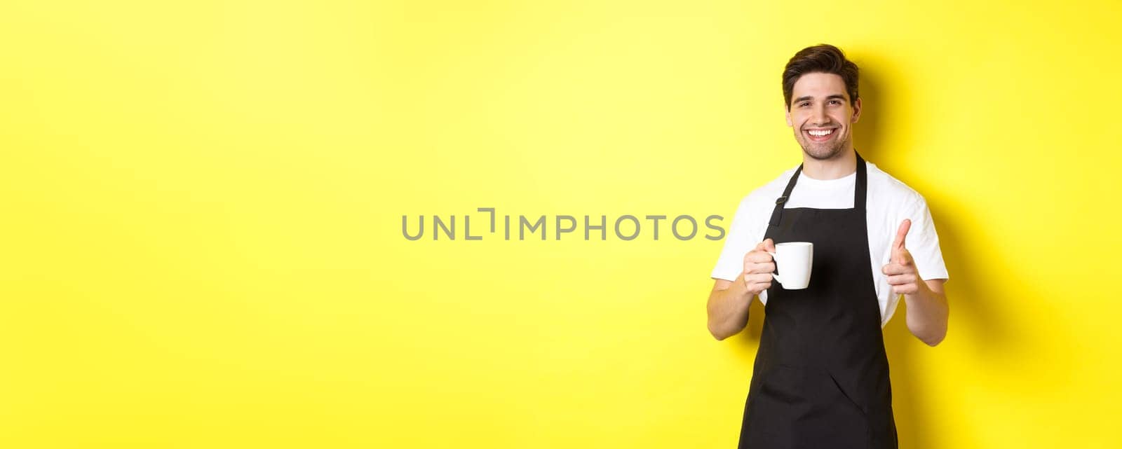 Barista bringing coffee and pointing finger gun at camera, standing in black apron against yellow background.