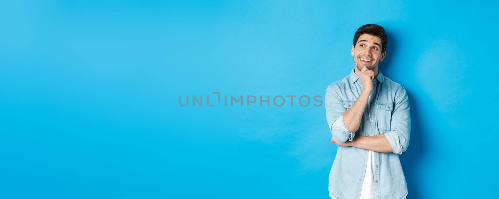 Portrait of thoughtful handsome man with beard, standing in casual outfit, looking at upper left corner and smiling, imaging or dreaming about something, standing over blue background.
