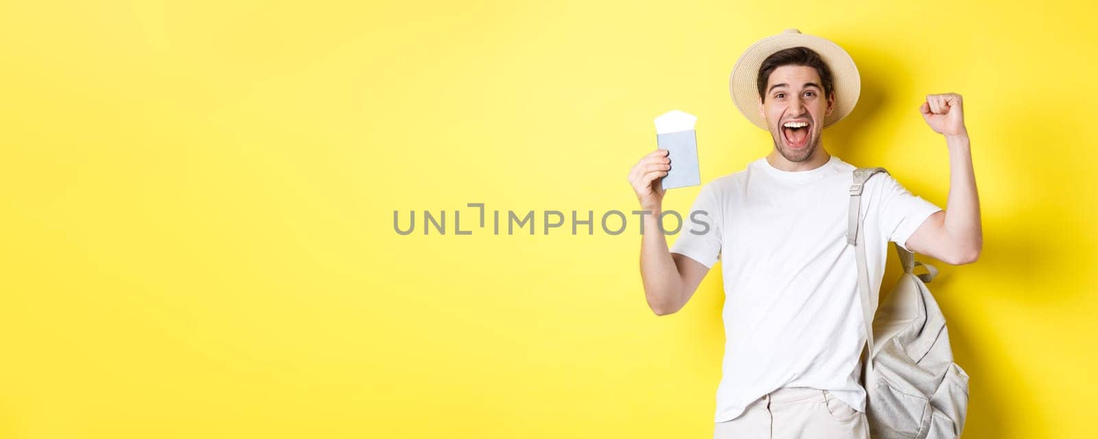 Tourism and vacation. Man feeling happy about summer trip, holding passport with plane tickets and backpack, raising hands up in celebration gesture, yellow background.