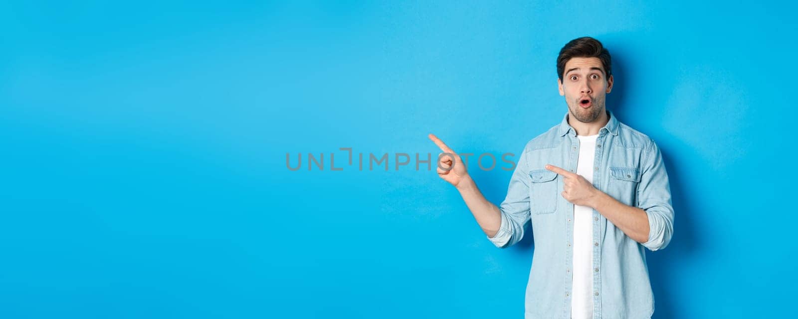 Portrait of surprised adult man in casual clothes showing announcement, pointing fingers left and looking amazed, standing against blue background.