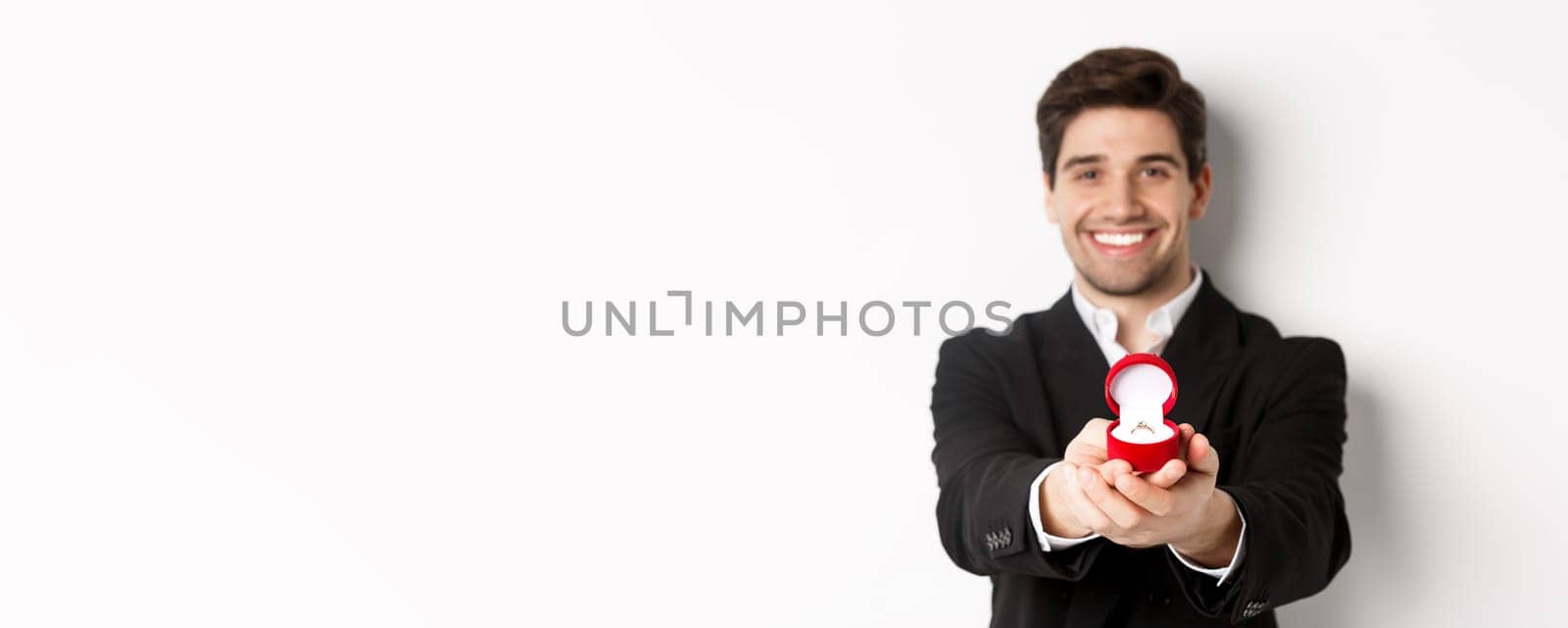 Image of handsome man looking romantic, giving you an engagement ring, making proposal to marry him, standing against white background.