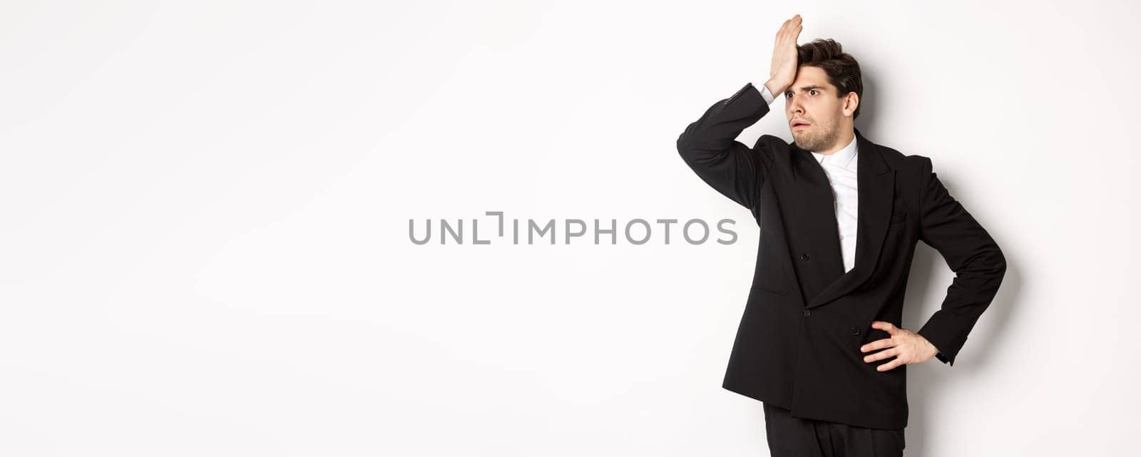 Portrait of shocked and distressed businessman in black suit, slap forehead and looking anxious, standing troubled over white background.