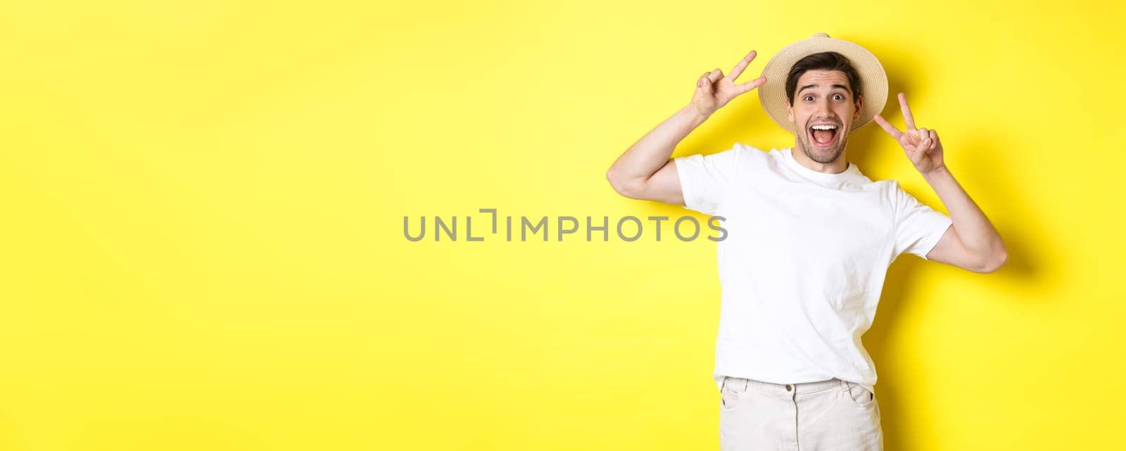 Concept of tourism and vacation. Happy male tourist posing for photo with peace signs, smiling excited, standing against yellow background.