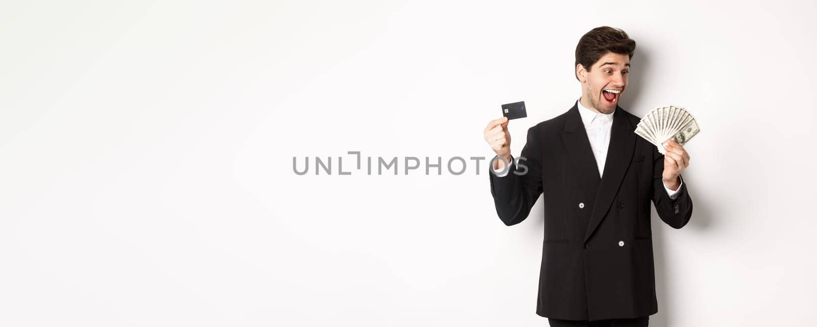 Image of attractive businessman in black suit, rejoicing, showing credit card and looking at money, standing against white background.