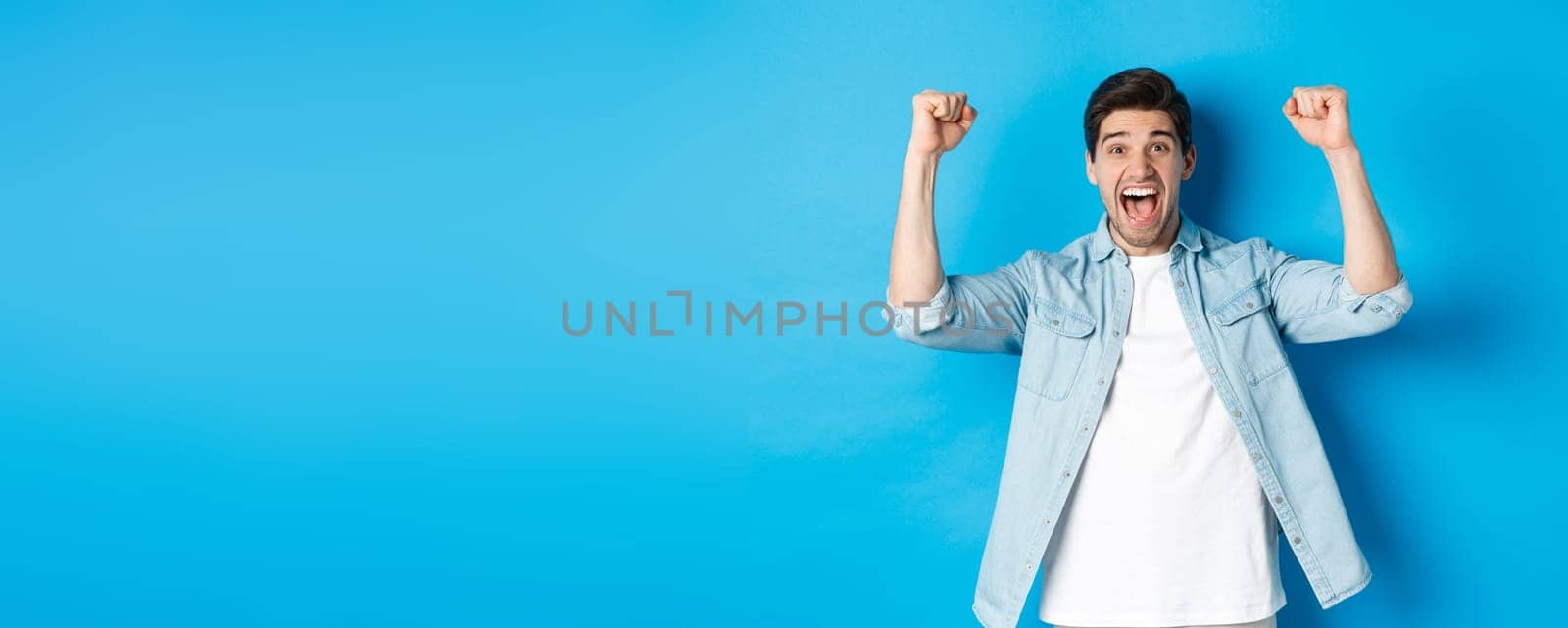 Happy attractive man triumphing, screaming yes and raising hands up to celebrate win, achieve goal, standing against blue background by Benzoix