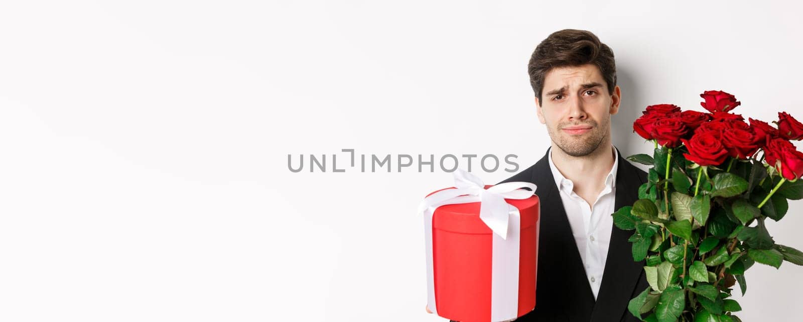 Close-up of sad man in suit, holding bouquet of red roses and a gift, standing upset against white background.