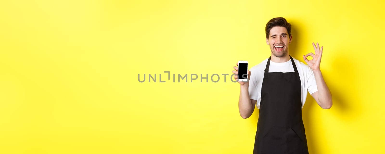 Handsome coffee shop worker showing ok sign and smartphone screen, recommending application, standing over yellow background.