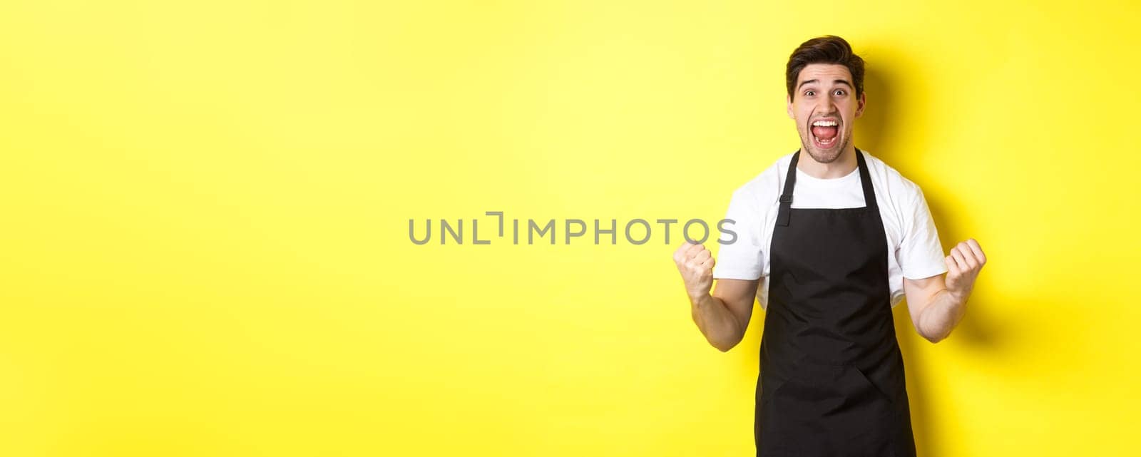 Excited coffee shop owner in black apron celebrating, making fist pump and shouting for joy, achieve goal, standing against yellow background.