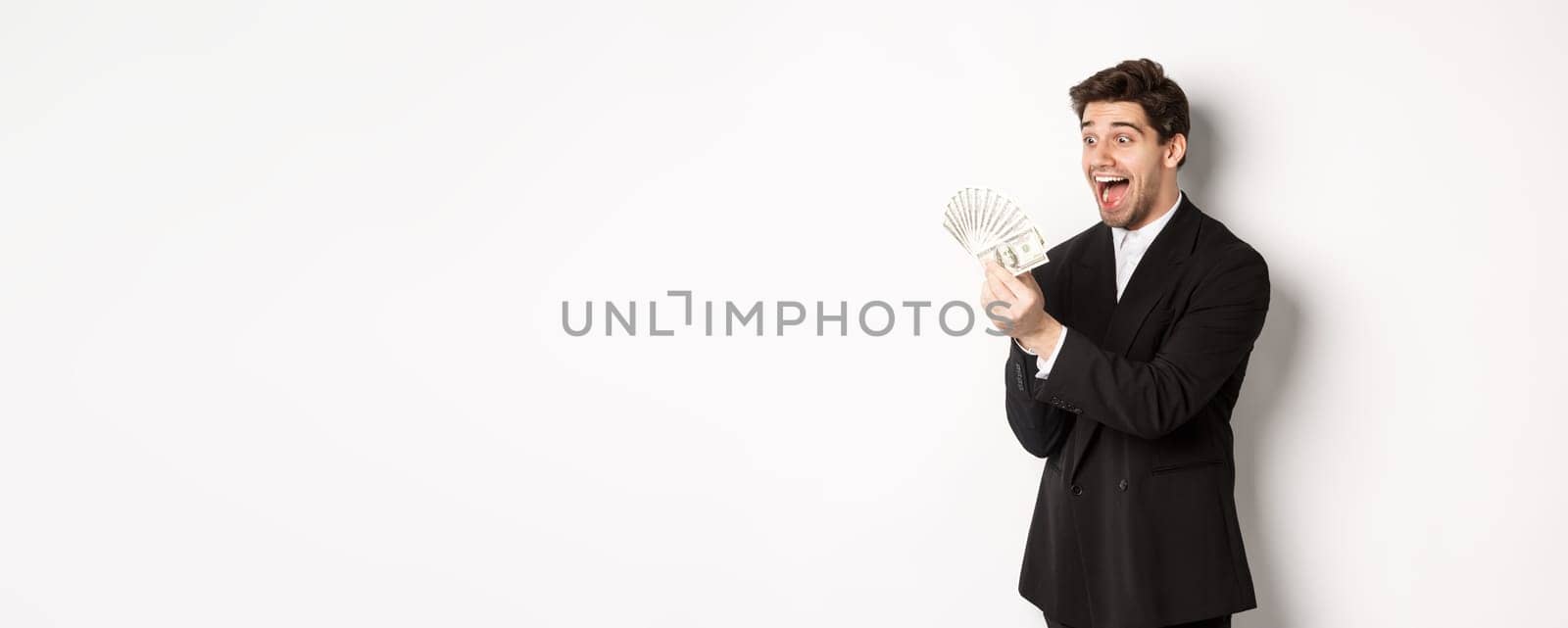 Image of excited handsome businessman in suit, looking amazed at money, winning cash, standing over white background by Benzoix