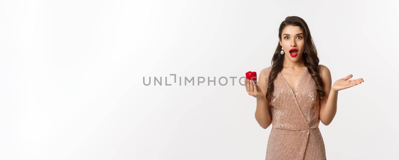 Image of amazed girlfriend holding engagement ring box, looking surprised, receive marriage proposal, wearing elegant dress, white background.