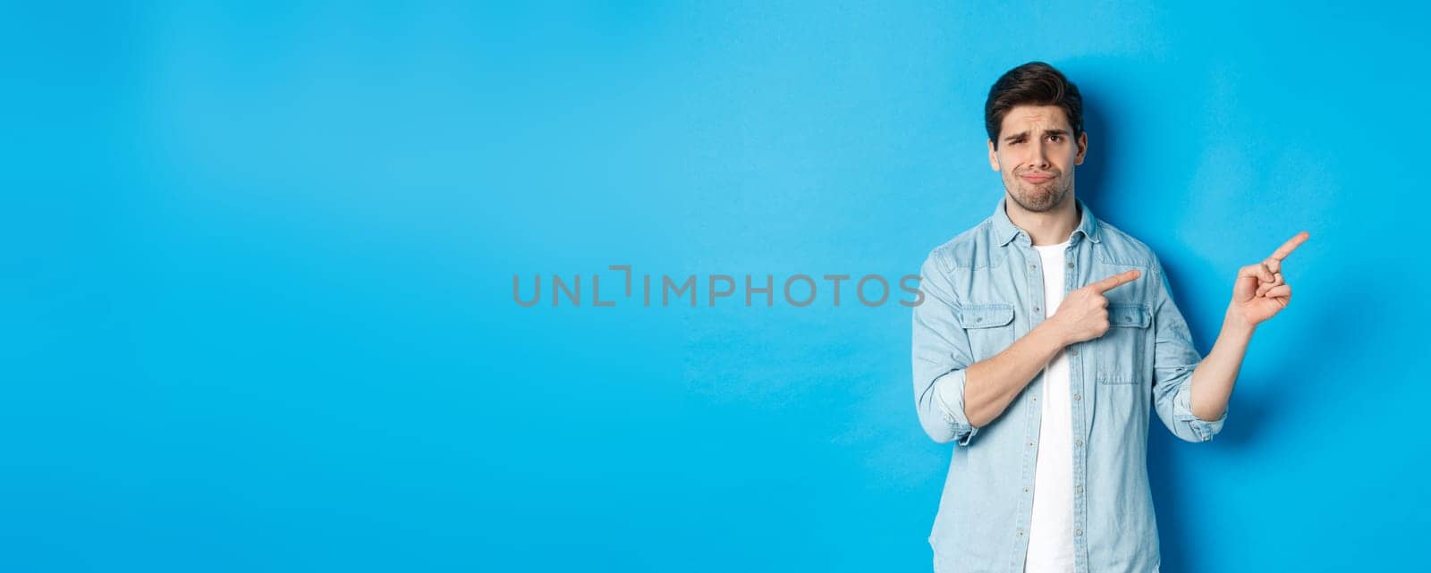 Portrait of skeptical adult guy pointing fingers right and smirking, exress disappointment and doubt, standing against blue background.
