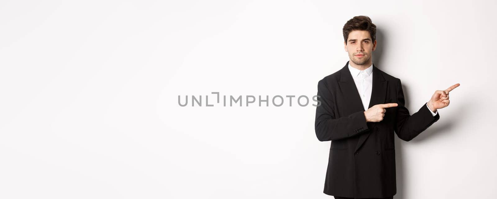 Image of handsome businessman in black suit, pointing fingers right and looking at camera, standing against white background.