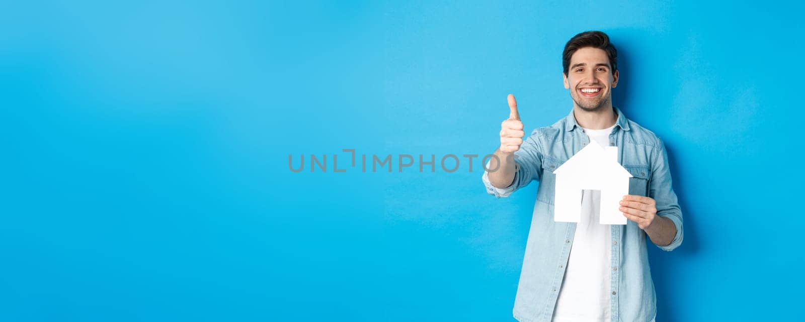 Insurance, mortgage and real estate concept. Satisfied client showing house model and thumb up, smiling pleased, standing against blue background.