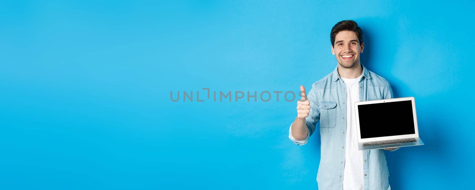 Smiling handsome man showing laptop screen and thumb-up, like promo offer, recommending website, standing against blue background.