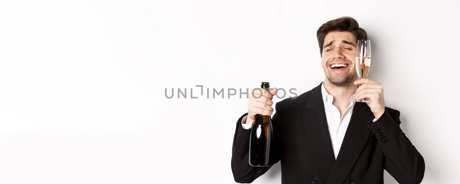 Close-up of handsome drunk guy in suit, holding glass of champagne and celebrating new year, standing over white background by Benzoix