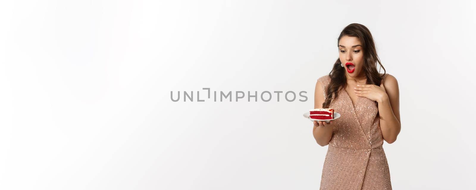 Party and celebration concept. Image of amazed slim female model in luxury dress, looking at piece of cake excited, standing over white background.